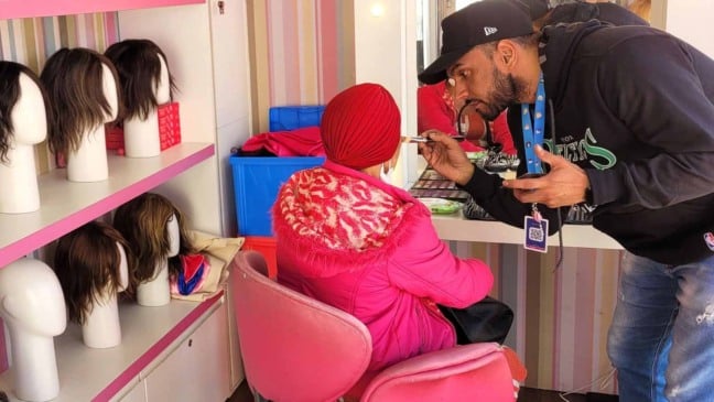 Profissional maquiador atende paciente oncológica usando lenço vermelho, em espaço com prateleiras cheias de perucas da ONG Cabelegria.