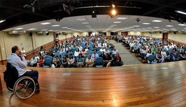 Cadeirante em palco falando para pessoas sentadas em cadeiras de auditório