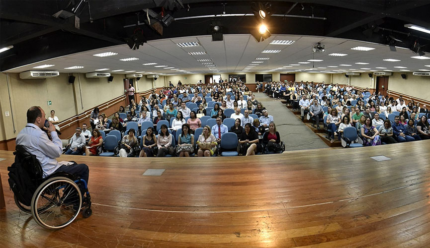 Cadeirante em palco falando para pessoas sentadas em cadeiras de auditório