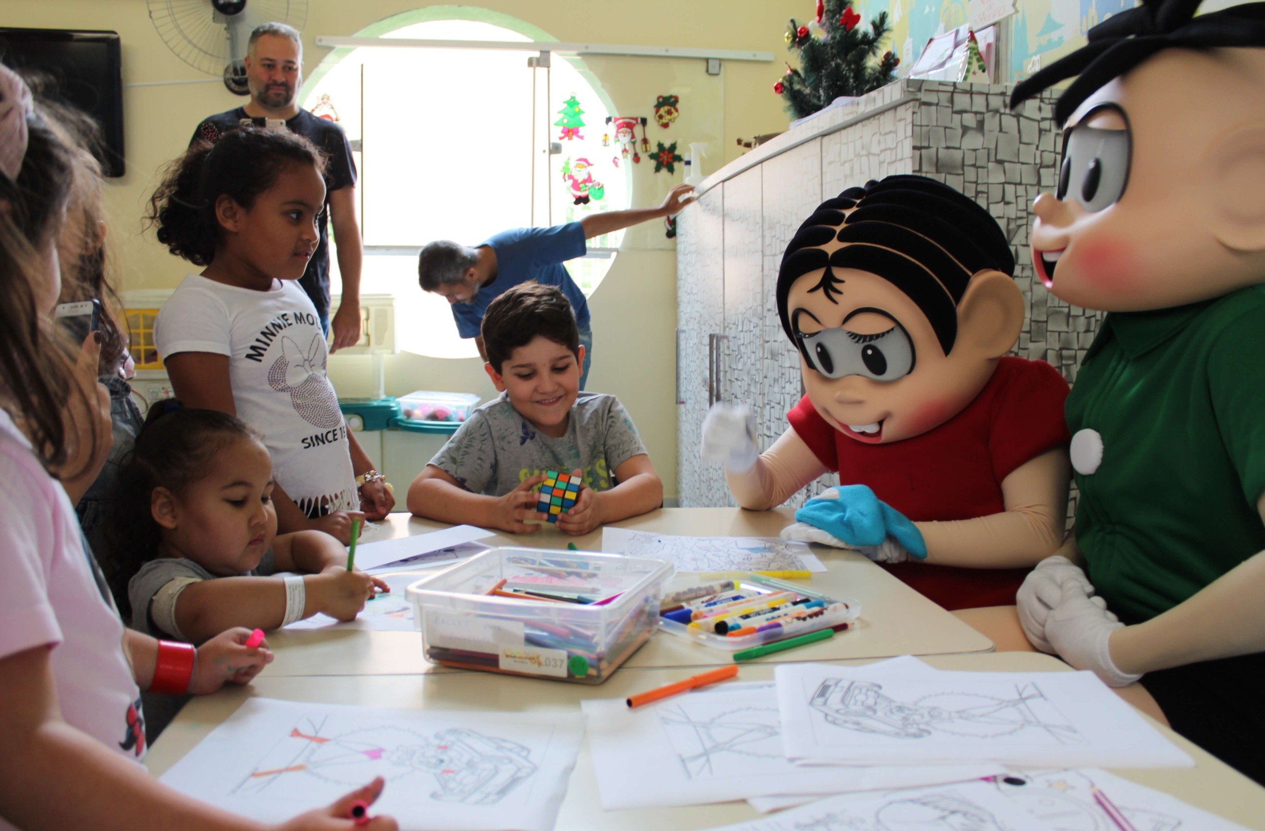 Mônica e Cebolinhas, personagens de Mauricio de Sousa, sentados com crianças em atividades de pinturas