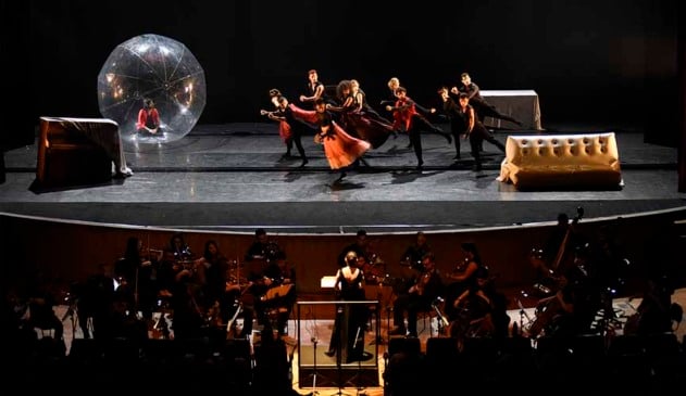 dançarinos em palco fazem coreografia, enquanto palco tem alguns itens de cenário. há bailarinos homens, com calça, e bailarinas mulheres, com as saias esvoaçantes. abaixo do palco, há um espaço onde uma orquestra segue os comandos de uma maestra.