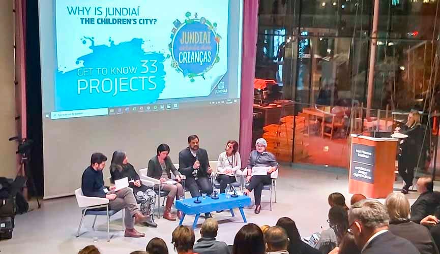 Prefeito de Jundiaí participando da abertura de evento na Holanda. Ao lado dele, outras pessoas sentadas, com imagem projetada atrás escrito em ingles "Porque Jundiaí é a Cidade das Crianças?". À frente, pessoas assistem a apresentação