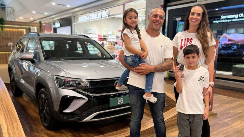 A imagem mostra uma família de quatro pessoas posando ao lado de um carro novo em um shopping. O grupo inclui um homem, uma mulher, uma menina e um menino. A imagem mostra uma família de quatro pessoas posando ao lado de um carro novo em um shopping. O grupo inclui um homem, uma mulher, uma menina e um menino.