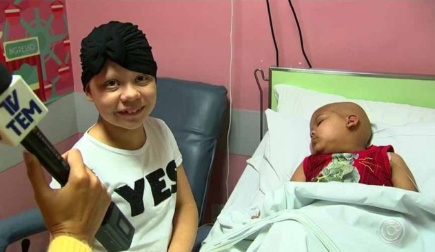 Larissa, menina com doença rara que causa queda dos pelos do cabelo, posando ao lado do pequeno Enzo, de 5 anos, internado por leucemia. Enzo está deitado na maca e dormindo.