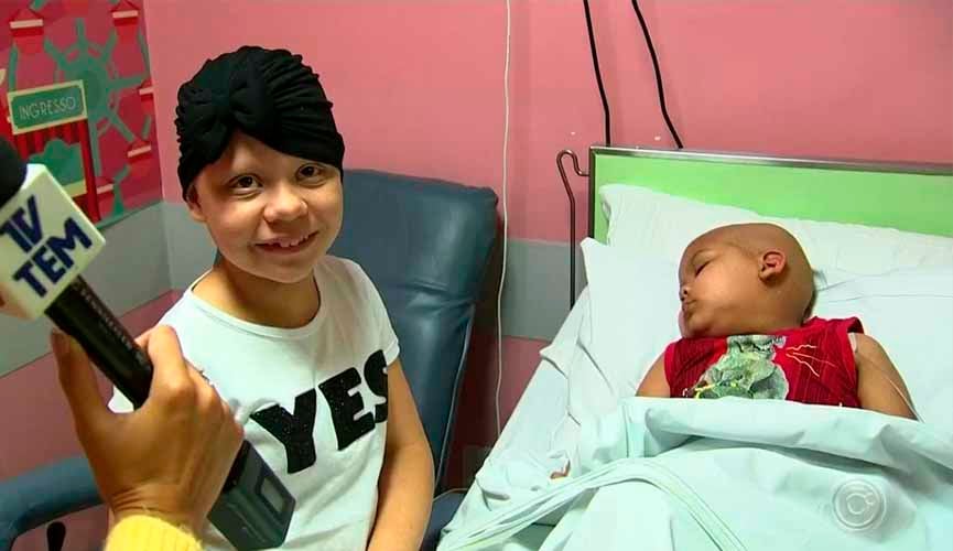 Larissa, menina com doença rara que causa queda dos pelos do cabelo, posando ao lado do pequeno Enzo, de 5 anos, internado por leucemia. Enzo está deitado na maca e dormindo.