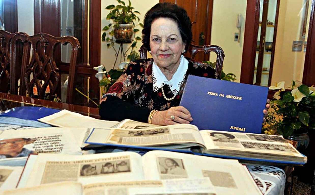 Imagem de Mercedes Ladeira Marchi, fundadora da Feira da Amizade, segurando um álbum azul com o título "Feira da Amizade Jundiaí