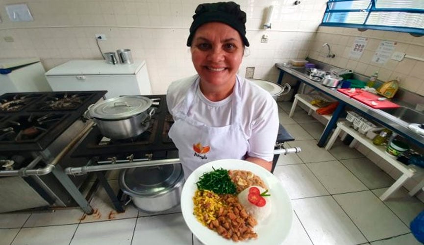Merendeira em cozinha segurando prato com arroz, pernil acebolado, tutu de feijão, farofa e salada de couve