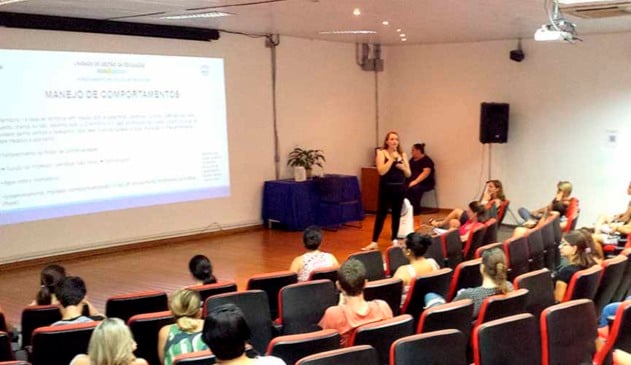 professora à frente de público, em cima de palco, fala ao microfone com slide na parede atrás. outros professores estão sentados nas cadeiras do auditório assistindo
