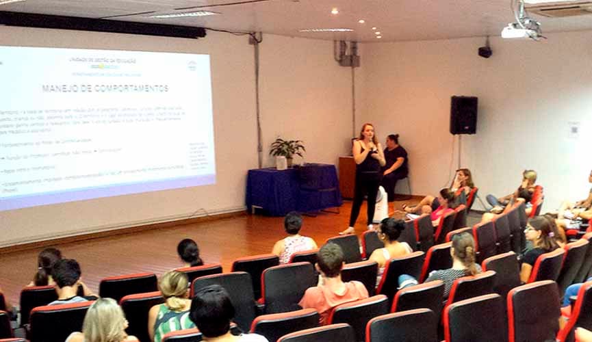 professora à frente de público, em cima de palco, fala ao microfone com slide na parede atrás. outros professores estão sentados nas cadeiras do auditório assistindo
