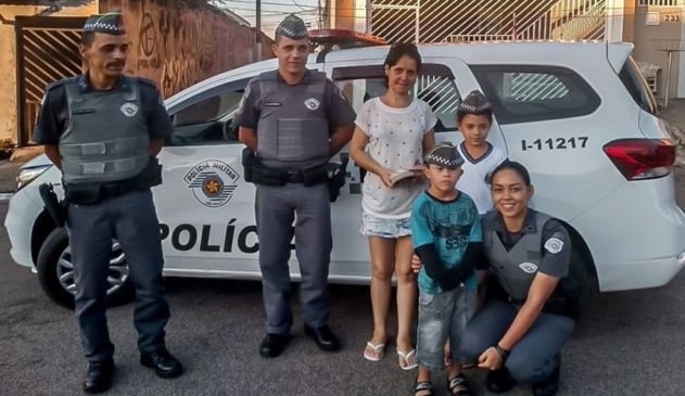 Policiais militares ao lado de menino com síndrome de Down e mãe de menino; ao fundo uma viatura da corporação e casas.