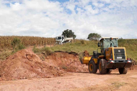 Trator realizando terraplanagem em uma área rural, com um caminhão ao fundo e uma plantação ao lado.
