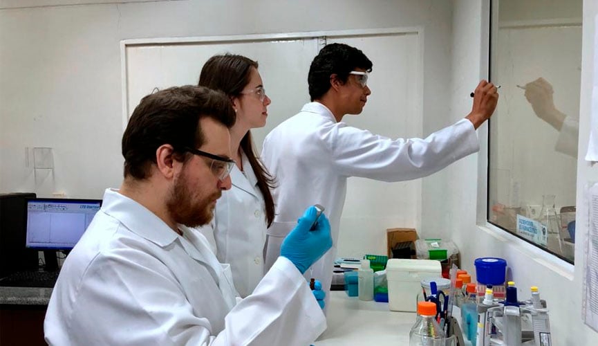 Três pesquisadores da Unicamp, sendo um á frente, analisando um material, enquanto uma mulher e um homem ao fundo analisam escritos na lousa