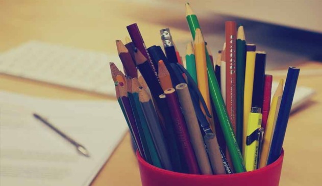 Porta lápis em destaque com variados lápis, de várias cores. ao fundo, desfocado, um papel com uma caneta em cima