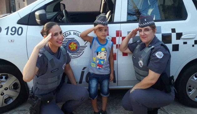 policiais militares mulheres agaixadas em frente à viatura. ao meio, há um menino de 5 anos, fazendo sinal de marcha como elas e com chapéu de policial