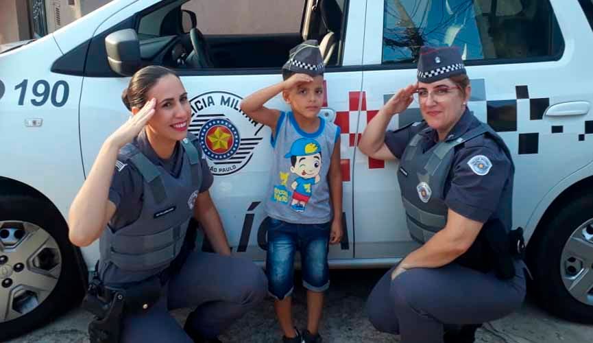 policiais militares mulheres agaixadas em frente à viatura. ao meio, há um menino de 5 anos, fazendo sinal de marcha como elas e com chapéu de policial