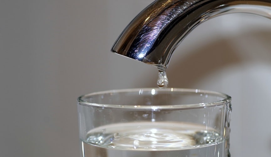 Copo cheio de água em baixo de torneira em que um último pingo de água cai