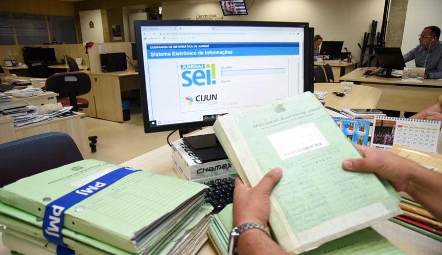 mãos de pessoa segurando uma pasta com uma série de documentos em papel, ao lado de outras pastas. à frente, monitor de computador mostra página aberta do novo sistema da prefeitura que moderniza e coloca no âmbito digital todos os processos internos de gestão