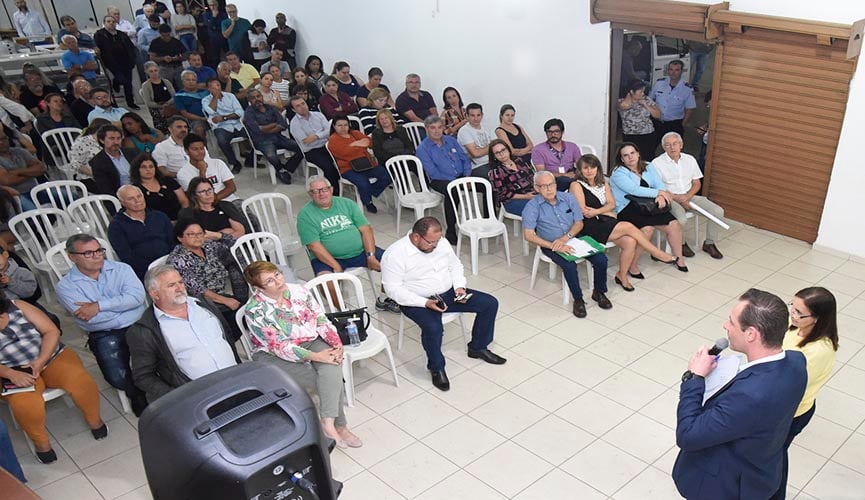 Pessoas sentadas em cadeiras de plástico em salão ouvindo gestor da saúde de jundiaí, Tiago Texera, falar ao microfome