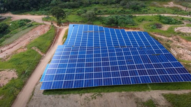 Na foto, vemos uma usina fotovoltaica em construção. A imagem mostra uma grande área coberta por inúmeras placas solares organizadas de maneira ordenada. O local onde a usina está situada é uma região verde e aberta, com vegetação ao redor e algumas estradas de terra que circundam o terreno. As placas solares estão dispostas em linhas e colunas, ocupando um espaço considerável, indicando um projeto de grande escala. A usina está localizada em uma área rural, longe de grandes edificações, o que permite um aproveitamento eficiente da luz solar.