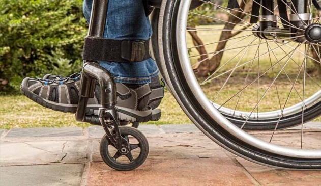 imagem da close em roda de cadeira de rodas e em perna de deficiente. foto é lateral e mostra pedaço da perna e do sapato, assim como pedaço da roda