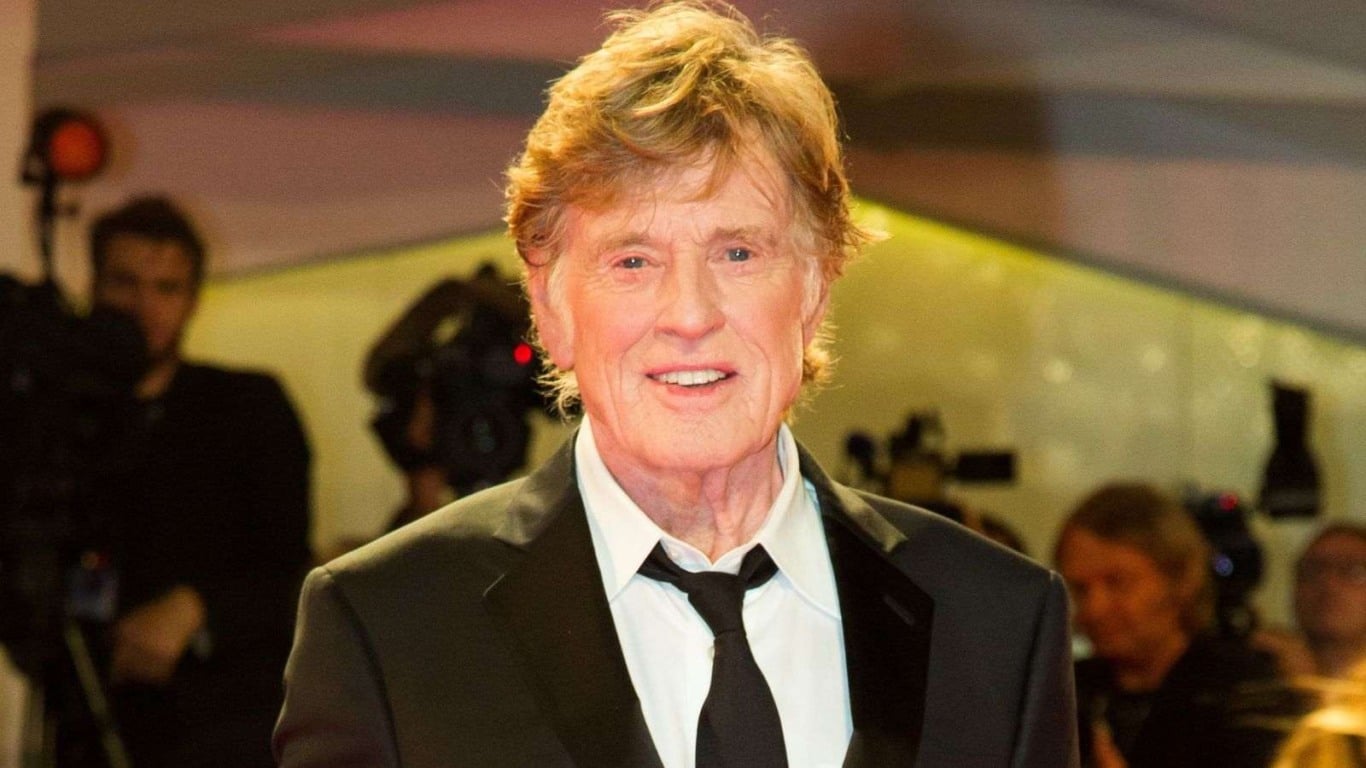 Robert Redford, ator e diretor vencedor do Oscar, em evento de gala usando terno preto e gravata, com câmeras e fotógrafos desfocados ao fundo.