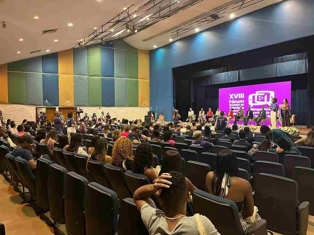 Auditório cheio durante o XVIII Fórum Brasileiro de Televisão Universitária, com palestrantes no palco e público atento.