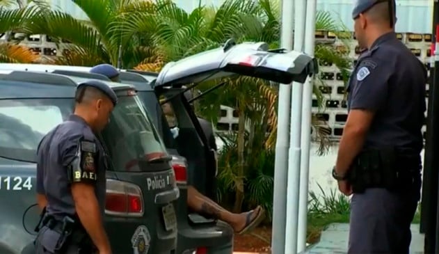 Reprodução de reportagem mostra policiais do lado de fora do caror e o porta-malas da viatura aberto com uma pessoa dentro. A perna da pessoa está para fora e visível