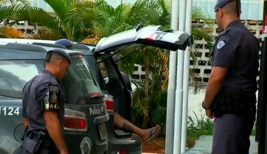 Reprodução de reportagem mostra policiais do lado de fora do caror e o porta-malas da viatura aberto com uma pessoa dentro. A perna da pessoa está para fora e visível