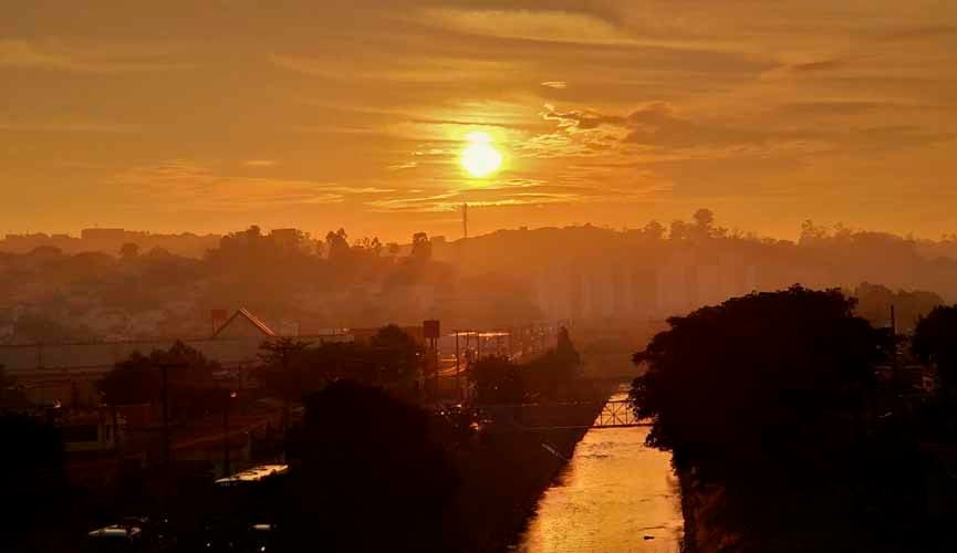 amanhecer com o sol alaranjado e silhuetas de prédios ao fundo. no meio da imagem corre um rio