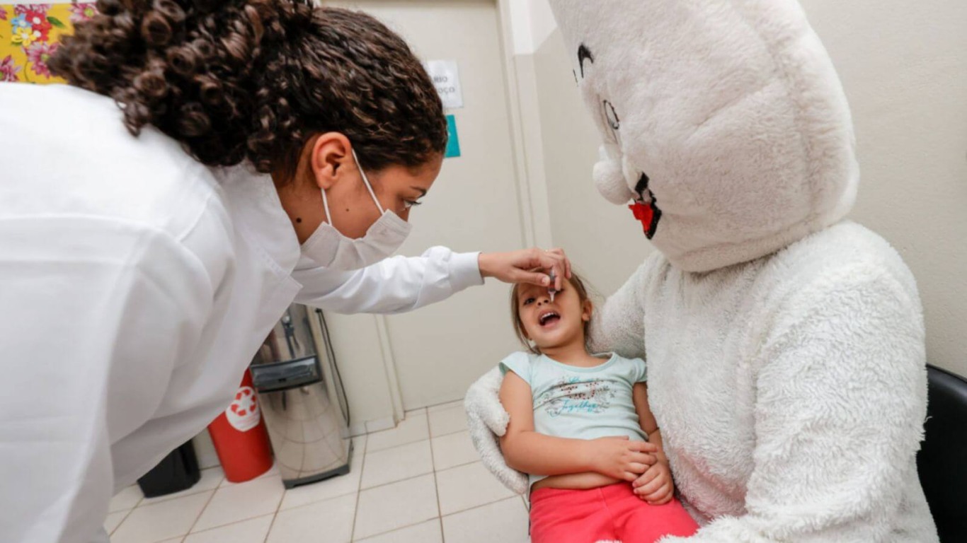 Criança deitada em colo de mascote do Zé Gotinha recebendo vacinação contra paralisia infantil