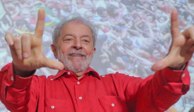 lula faz sinal de L com as duas mãos, enquanto sorri e veste uma camisa vermelha