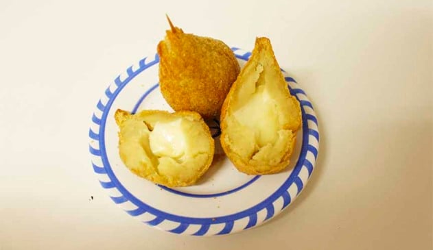 Duas coxinhas de queijo dentro de um prato. Uma delas dividida ao meio, com as duas partes deitadas e a outra, inteira, com um palito fincado na ponta.