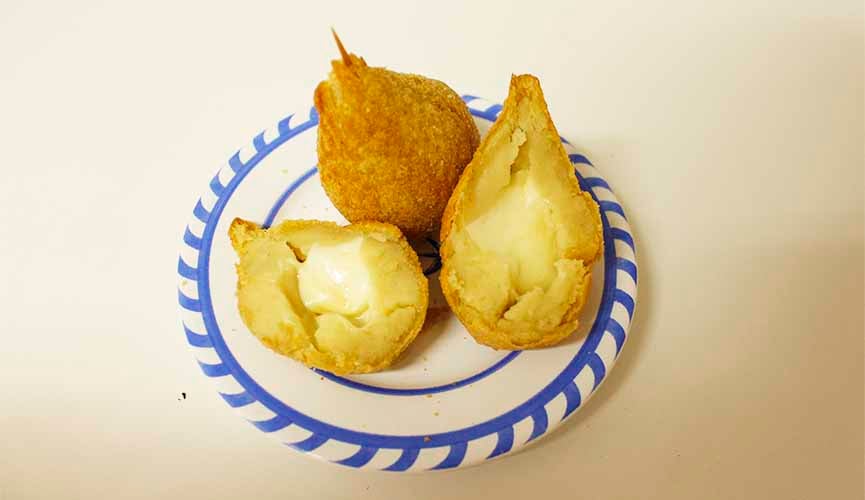 Duas coxinhas de queijo dentro de um prato. Uma delas dividida ao meio, com as duas partes deitadas e a outra, inteira, com um palito fincado na ponta.