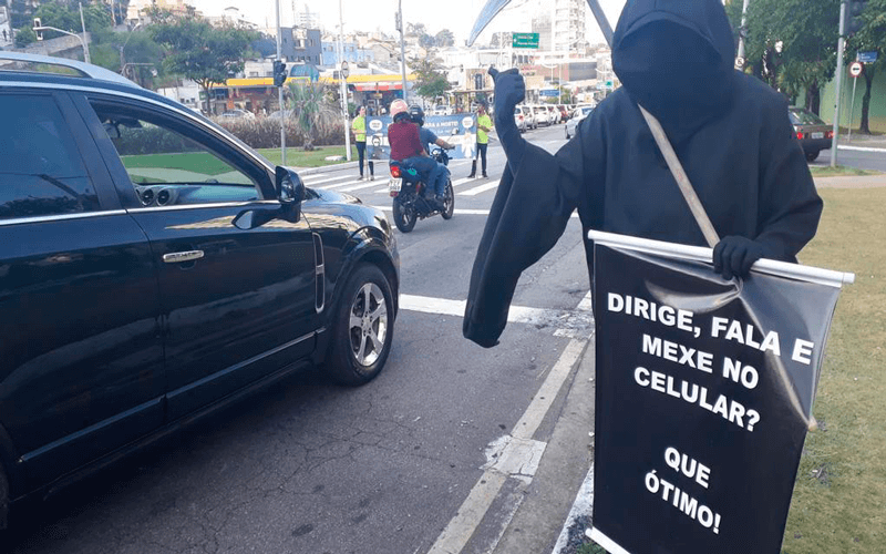 Ator vestido de morte, com capa preta e foice, exibe um cartaz na Av. 9 de Julho: “Dirige, fala e mexe no celular? Que ótimo!”