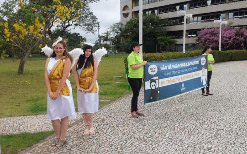 Duas atrizes vestidas de anjos estão à frente, enquanto dois membros do Unidade de Mobilidade e Transporte de Jundiaí estão atrás, exibindo um banner de conscientização.