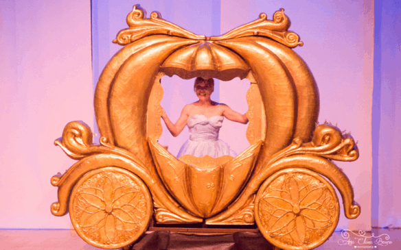 Foto de trecho do espetáculo em que Cinderela está dentro de uma carruagem que se assemelha a uma grande abóbora.