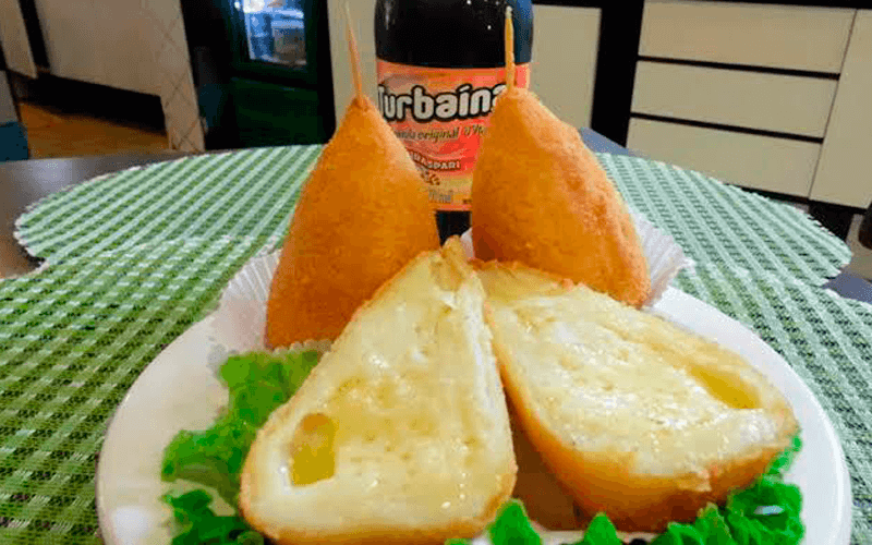 Coxinha de queijo cortada ao meio, com garrafa de Turbaína atrás.