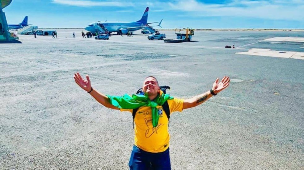 Foto de Anderson ajoelhado em um pátio de aviões vestindo camisa brasileira, com bandeiras enrolada no pescoço. Ele ergue as mãos para o céu como forma de agradecimento