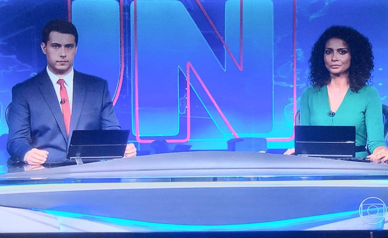 Giovanni à esquerda, sentado na bancada do Jornal Nacional, com a jornalista da noite ao lado