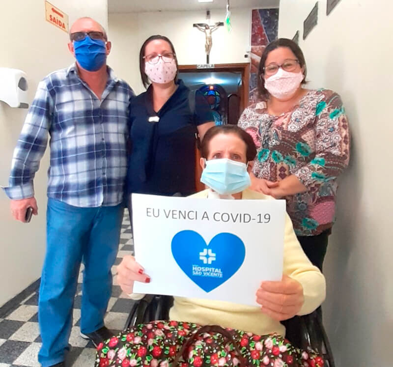 Maria Bueno de cadeira de rodas, ao lado da família, com cartaz: "eu venci a covid-19"