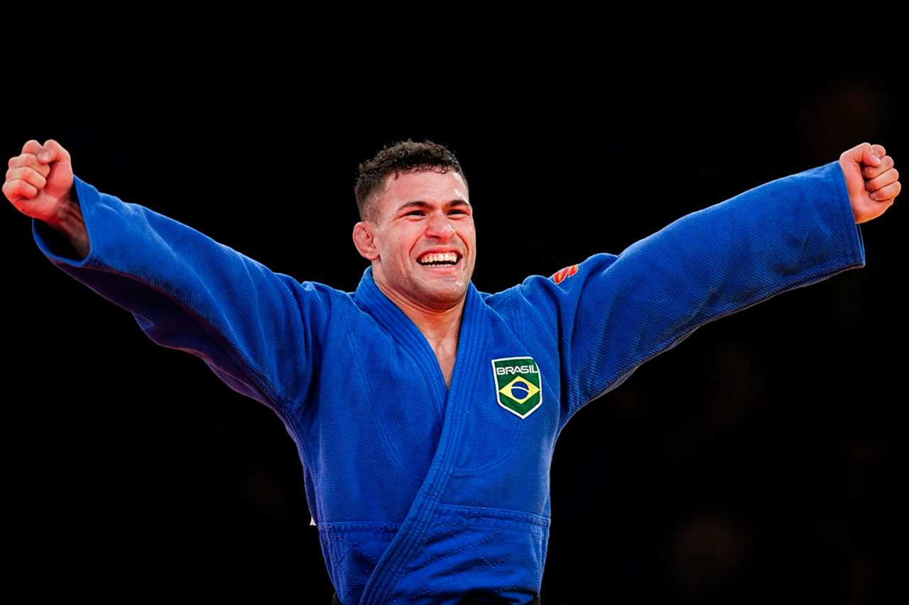 Judoca brasileiro comemorando vitória nos Jogos Olímpicos de Paris 2024, vestindo kimono azul e sorrindo com os braços levantados.
