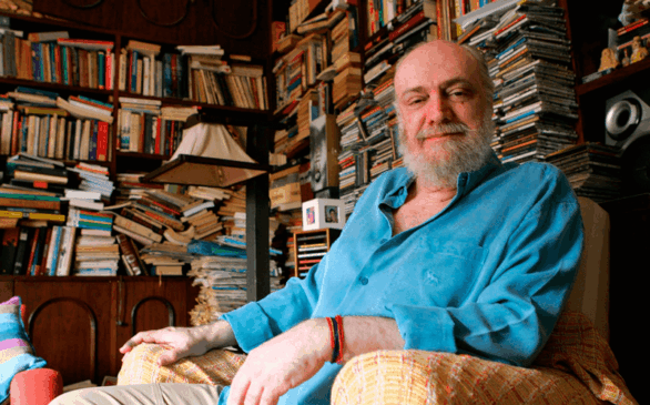 Aldir Blanc sentado em poltrona de biblioteca