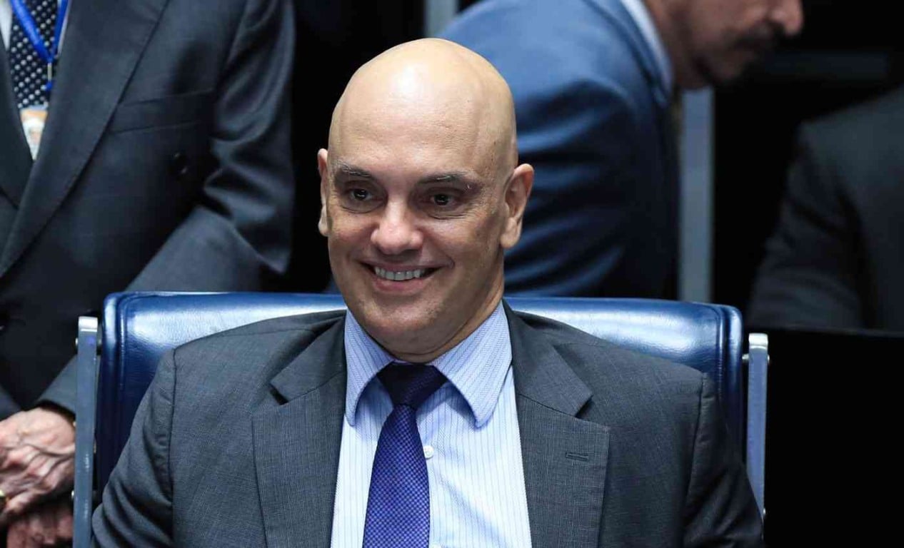 Ministro do STF Alexandre de Moraes de terno e gravata azul sorrindo, sentado em uma cadeira, com outras pessoas ao fundo em um ambiente formal.