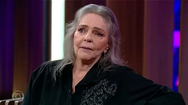 Angela Ro Ro em entrevista na TV, com expressão séria, usando camisa preta.