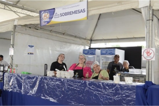 Voluntários sorridentes atendem barraca de sobremesas em evento dos 60 anos de Várzea Paulista, com banner e refrigeradores ao fundo.