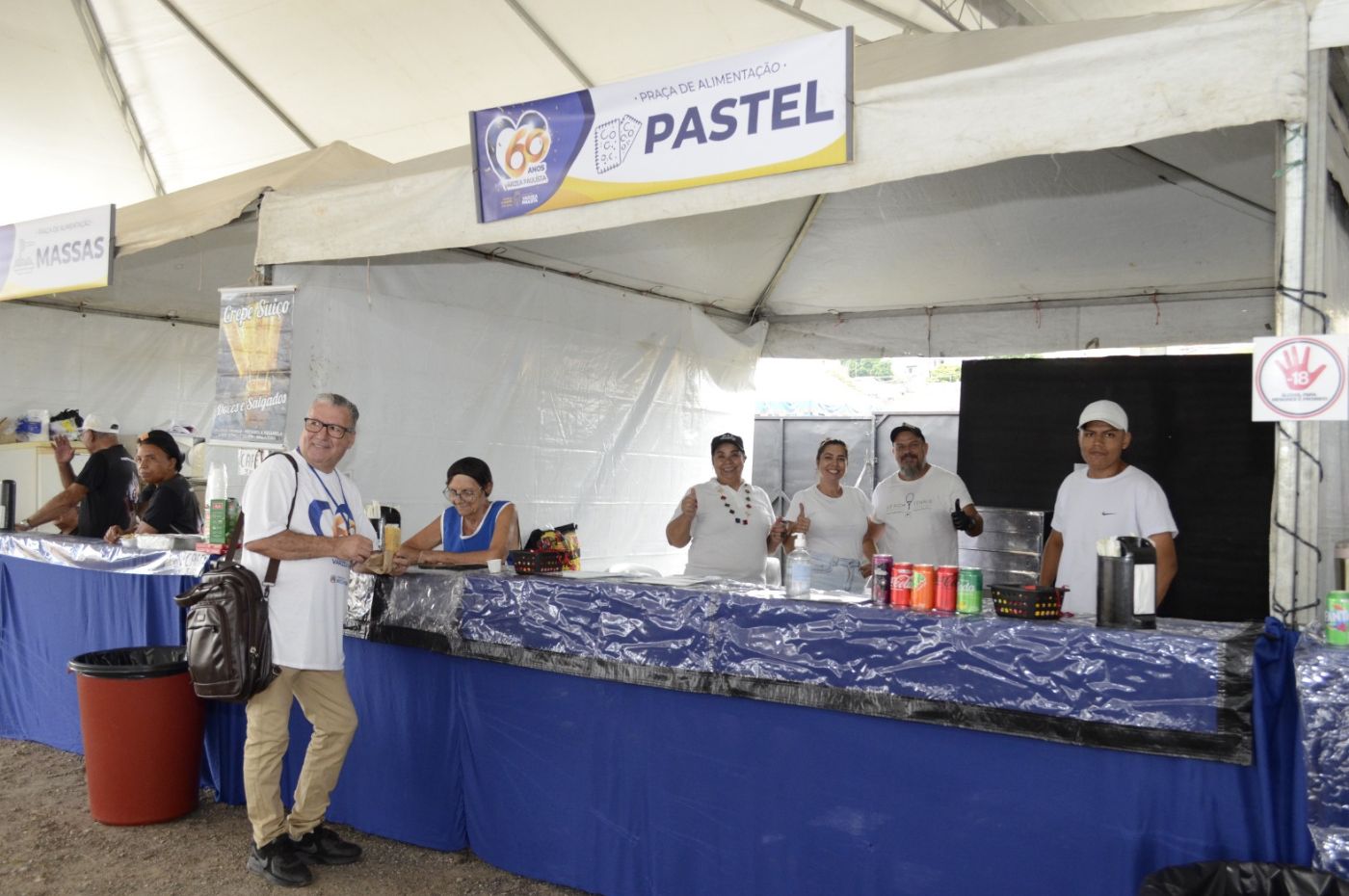 Voluntários servem pastéis em barraca do evento de 60 anos de Várzea Paulista, com clientes sorridentes e bebidas à mostra.