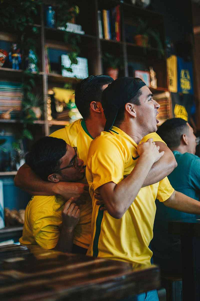 Três torcedores brasileiros comemorando em um bar enquanto assistem a um jogo, representando a emoção das apostas online. Três torcedores brasileiros comemorando em um bar enquanto assistem a um jogo, representando a emoção das apostas online.
