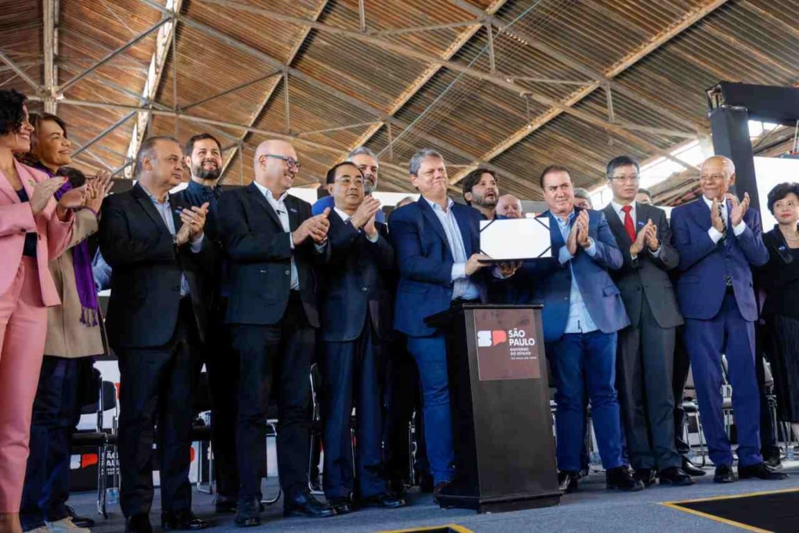 Pessoas de terno em um evento oficial, aplaudindo e segurando um documento, com o logotipo do Governo de São Paulo ao lado.