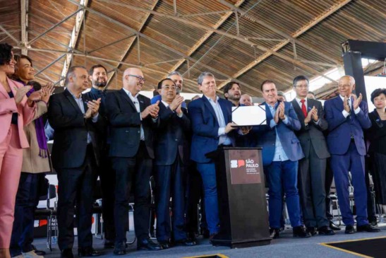 Pessoas de terno em um evento oficial, aplaudindo e segurando um documento, com o logotipo do Governo de São Paulo ao lado.