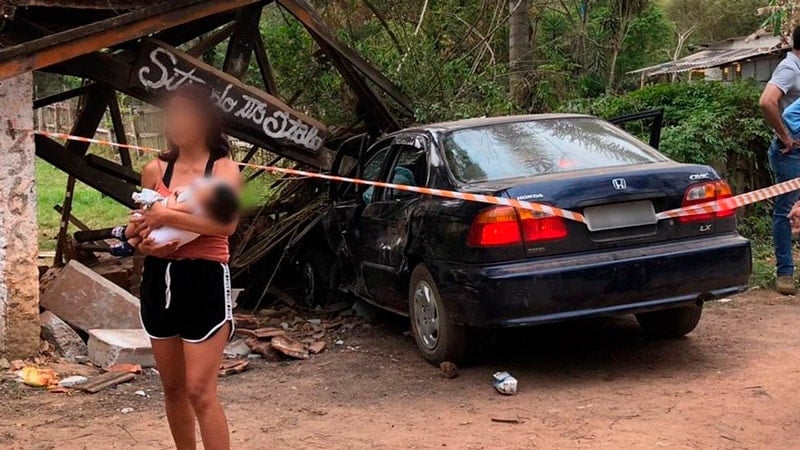 Carro destruído em ponto de ônibus; mulher com criança no colo